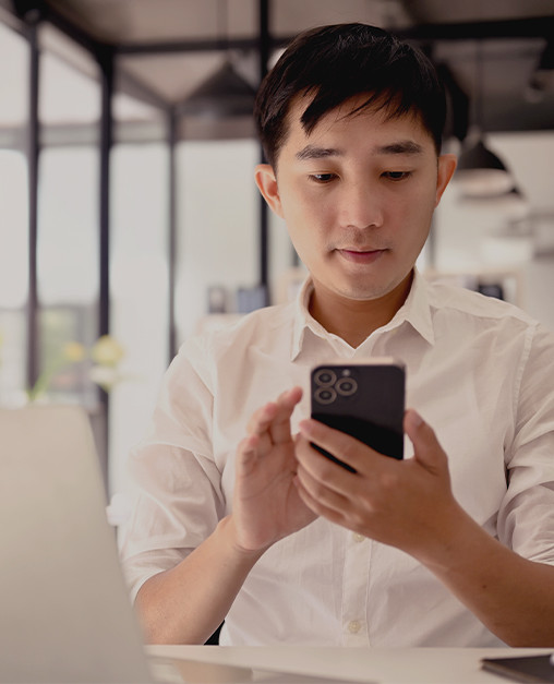 A young professional seated at a desk looks at a smartphone while a laptop is open in front of them, illustrating digital-first engagement alongside traditional work tools.
