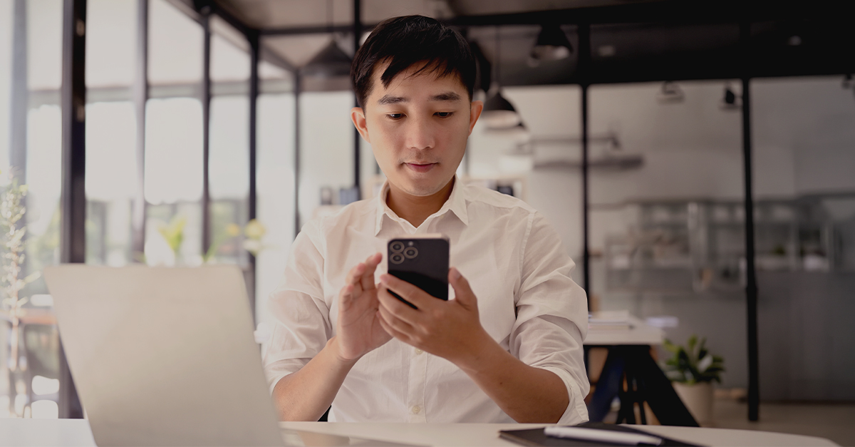 A young professional seated at a desk looks at a smartphone while a laptop is open in front of them, illustrating digital-first engagement alongside traditional work tools.