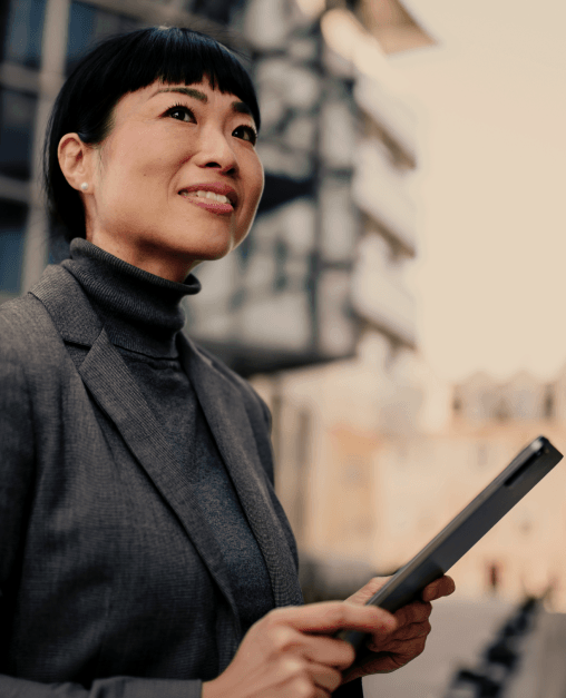 A person in professional clothing standing outdoors in an urban environment, holding a digital tablet.