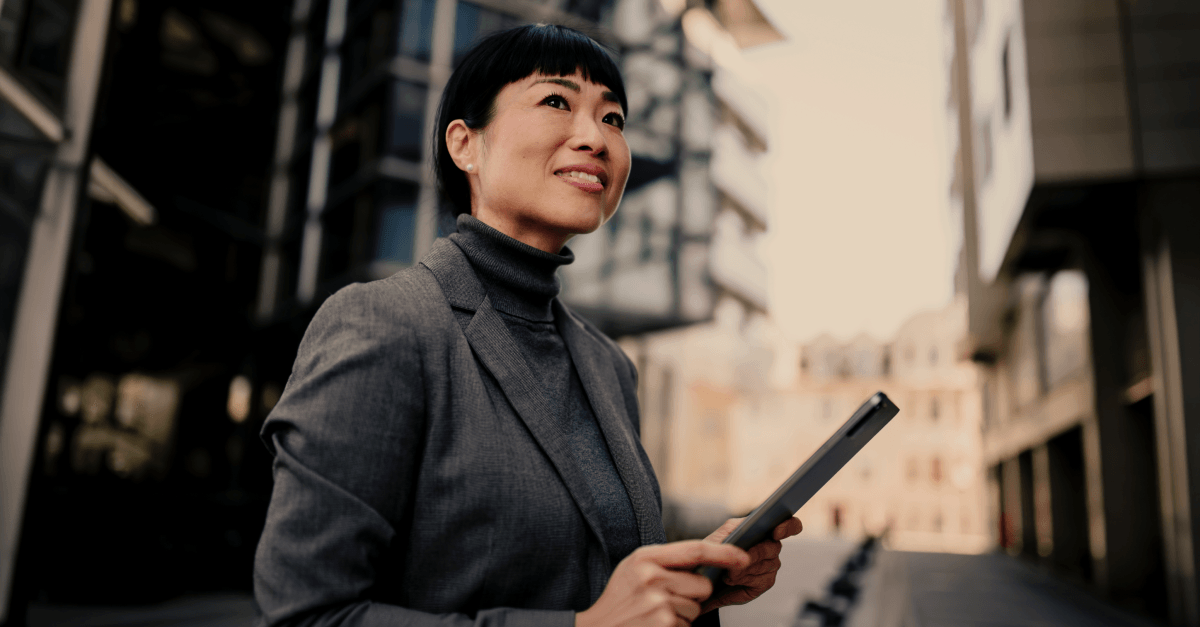 A person in professional clothing standing outdoors in an urban environment, holding a digital tablet.